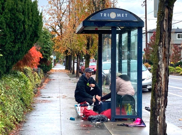 A Portland Street Response EMT crouches to assist an unhoused person who sits at a city bus stop with their possessions.